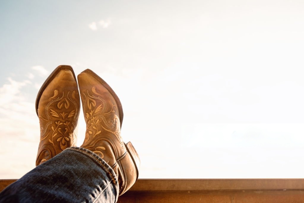 A close-up of a person wearing brown embroidered cowboy boots resting on a ledge under a bright sky, symbolizing Western style and relaxation.