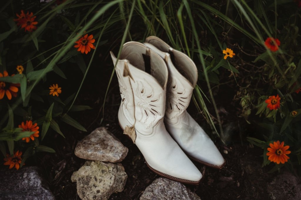 White cowboy boots placed on rocks surrounded by green grass and orange flowers.