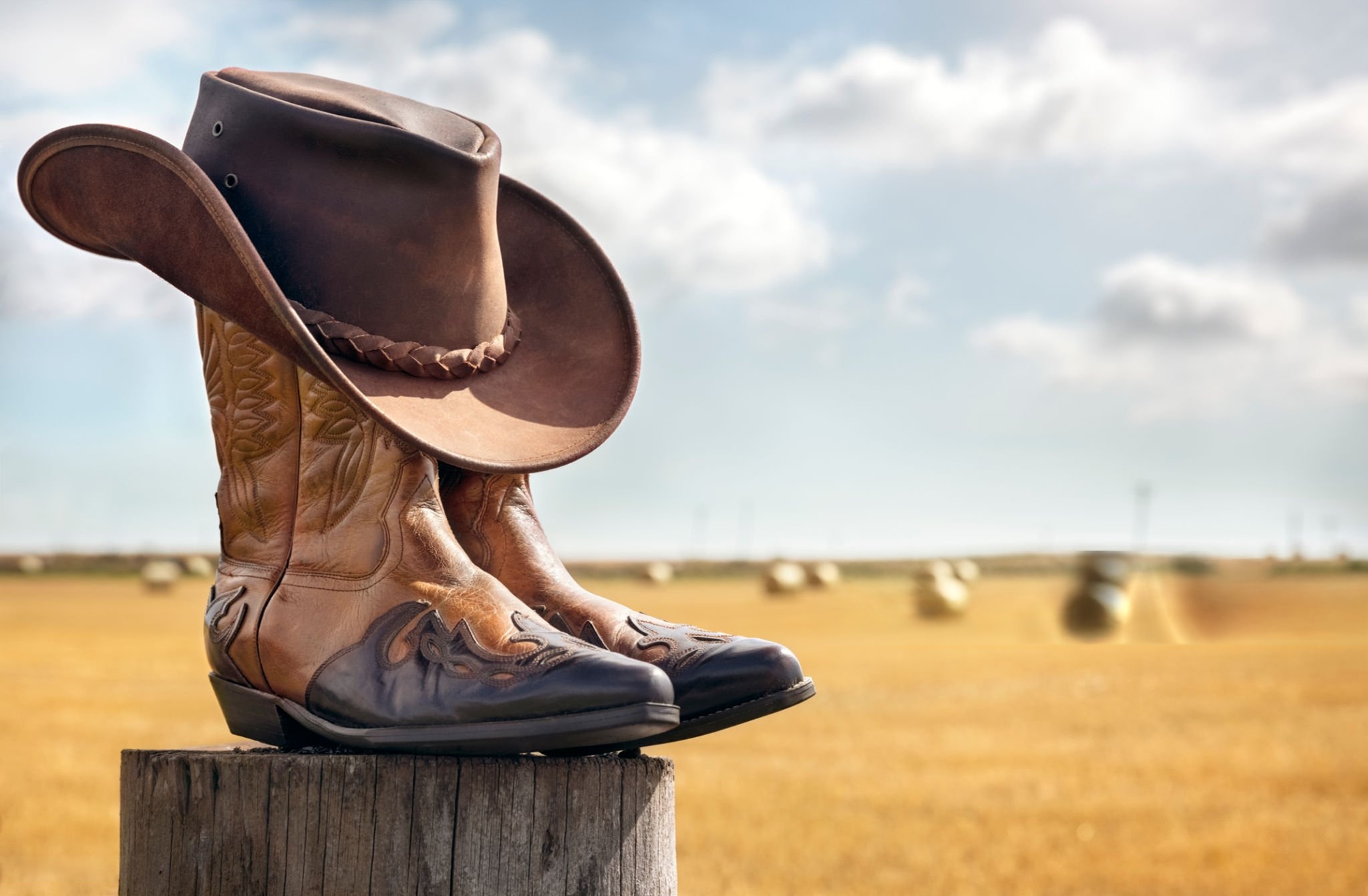 Feature image Cowboy hat resting on a pair of leather boots in a sunny field with hay bales in the background.